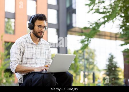 A young student wearing headphones works on his laptop outdoors on a sunny day, studying. Stock Photo