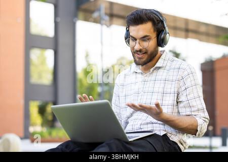 A young student wears headphones and engages in a video call outdoors, using his laptop with a friendly expression. Stock Photo