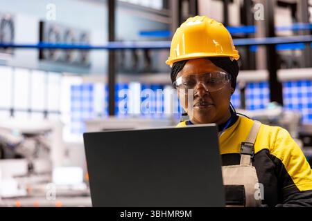 Closeup of female compliance engineer reviewing machinery software updates on her laptop. African american industrial technician working on developing innovative infrastructures in smart factory. Stock Photo