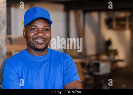African American man leaning on workshop column holding pencil behind ear near table saw and planks Stock Photo