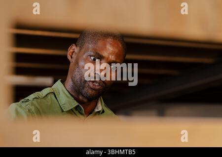 Mature African American man standing leaning forward in workshop examining wooden planks on bench Stock Photo