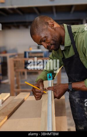 African American man carpenter measuring plank using tape measure and pencil in shop, copy space Stock Photo