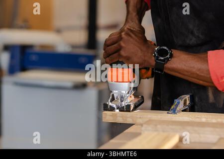 African American man operating jigsaw cutting wooden board secured by clamp at workshop, copy space Stock Photo