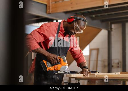 African American man cutting plank at woodshop bench wearing apron, goggles with orange jigsaw saw Stock Photo
