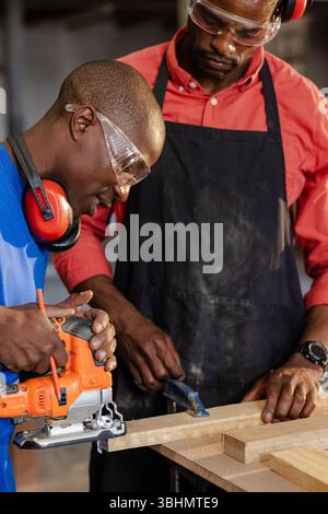African American male coworkers using jigsaw, holding clamp and cutting plank on workshop bench Stock Photo