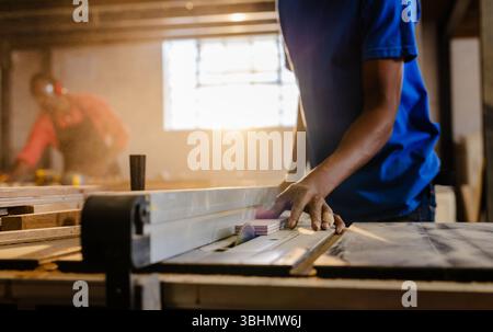 African American male coworkers operating table saw and sanding plank in dusty workshop, copy space Stock Photo
