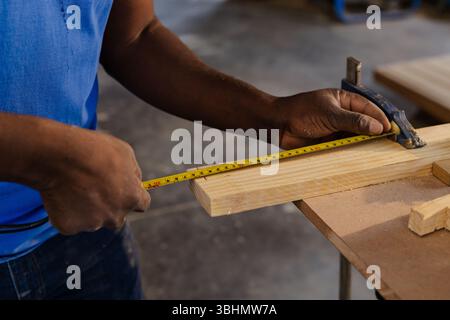 African American man measuring plank clamped to bench in carpentry shop using tape measure Stock Photo