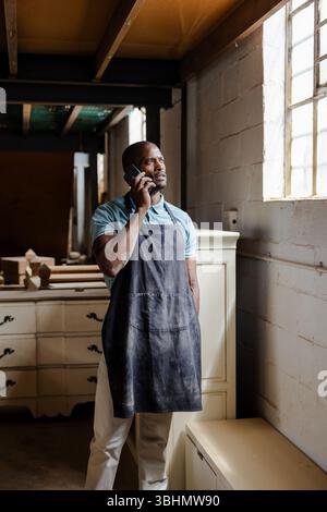 African American man wearing work apron checking smartphone near dressers in furniture workshop Stock Photo