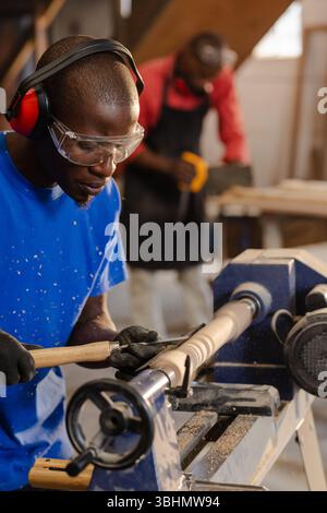 African American male coworkers shaping wooden spindle using lathe chisel and sanding wood at bench Stock Photo
