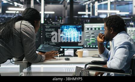 Admin in wheelchair and data center coworker working together using AI node tree software to minimize downtime. Server hub IT expert in disability and colleague using visual programming app, camera B Stock Photo