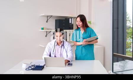 Asian adult doctor uses laptop at desk with nurse Asian adult reviewing patient chart in clinic. Medical professionals collaborating on healthcare. Pa Stock Photo