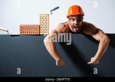 Crazy builder with bricks, funny sale on construction site. Funny man with helmet doing brick work. Builder posing with bricks blocks. Bricklayer Stock Photo