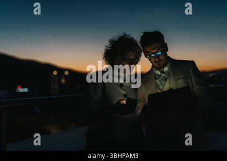 Two business people are focused and collaborating late at night, using digital devices outdoors. Stock Photo