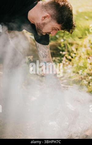 A man with tattoos on his arms is cooking food over a fire. The smoke from the fire is rising into the air, creating a hazy atmosphere. The man is foc Stock Photo