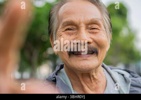 Head shot portrait of happy Asian senior man taking selfie, having fun with phone cam, smiling aged over nature background. Retirement lifestyle conce Stock Photo