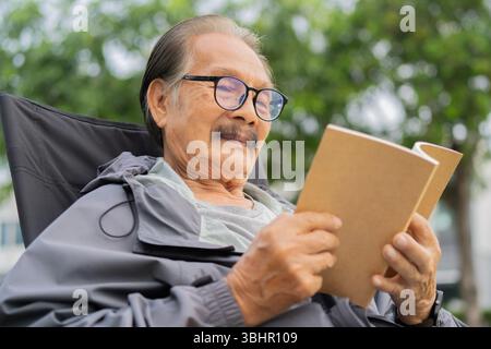 Side view portrait of elder Asian man sitting on an outdoor chair at nature and enjoying reading a book. Selective focus. Stock Photo
