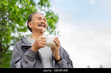 Handsome elderly man drinking, holding coffee from big cup while standing outdoor by the big tree in a summer day. Copy space. Stock Photo
