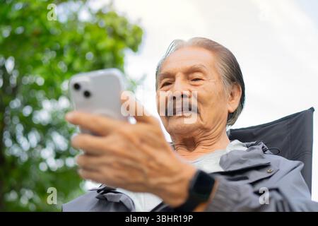Happy calm relaxed elderly man sitting on chair in city park enjoy with his smartphone. Selective focus. Elderly lifestyle concept. Stock Photo