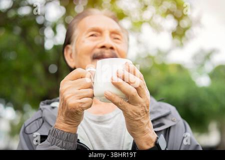 Healthy Asian senior man drinking coffee in morning park. Selective focus on hands. Getting cup of coffee concept. Stock Photo