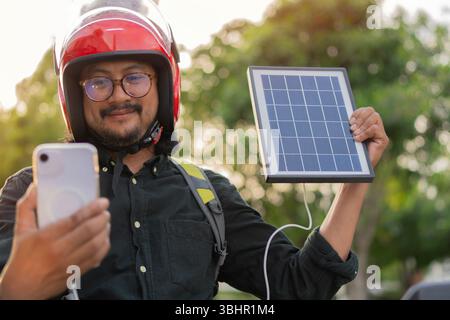Smiling handsome Asian man holding solar panel sitting on a motorcycle while using smart phone before leaving for work or travel. Alternative electric Stock Photo