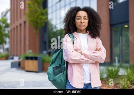A young black woman with curly hair poses confidently outdoors with a backpack, near a modern building, wearing a pink shirt. Stock Photo