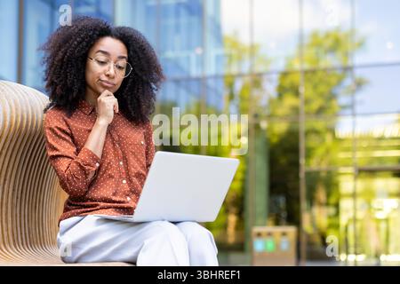 A thoughtful woman wearing glasses, working on a laptop outside. She is sitting and holding her chin. Stock Photo