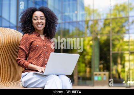 A beautiful, smiling woman with curly hair works on her laptop outdoors in front of a modern glass building. Stock Photo
