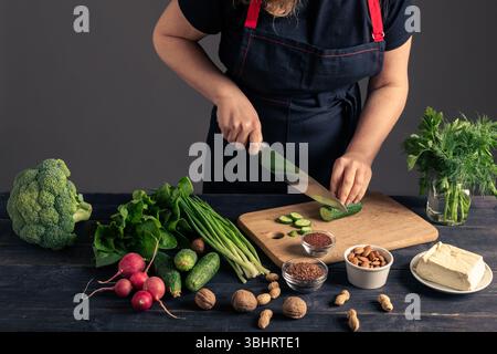 A woman in a black apron slicing fresh cucumber on a wooden cutting board. This image represents healthy eating, vegetarian lifestyle, and food prepar Stock Photo