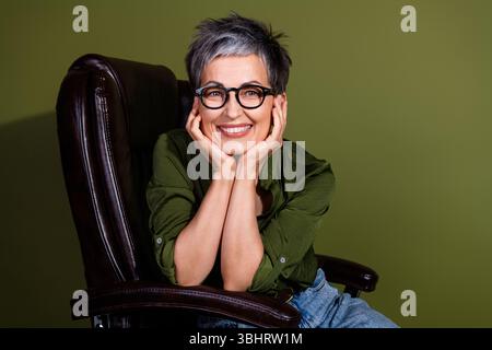Confident mature woman with short gray hair in glasses smiling warmly while posing casually against a khaki green background Stock Photo