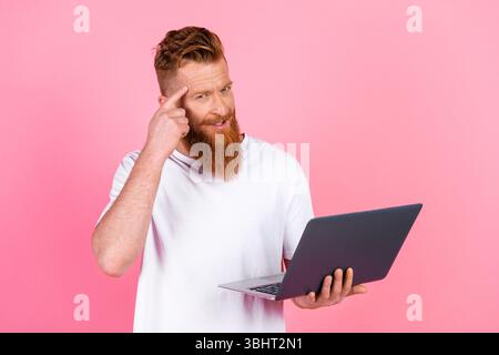 Young bearded man holding a laptop and thinking while standing against a pink background, showcasing casual style and leisure Stock Photo