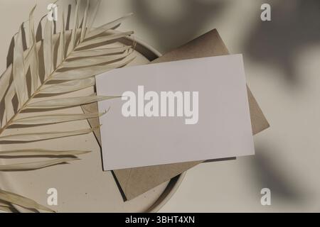 Blank horizontal greeting card, invitation mockup, craft envelope in sunlight, beige table background. Floral shadows overlay. Palm leaf on plate Stock Photo