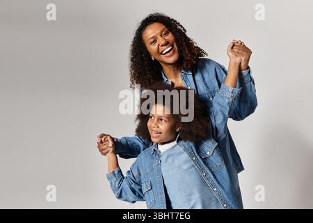 Smiling Black mother and daughter in denim, holding hands, grey background. Stock Photo