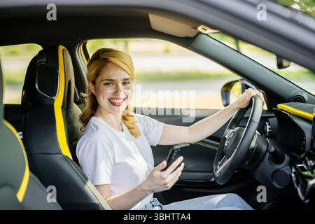 Happy woman demonstrates smartphone with white screen while sitting in car. Smiling woman showing a place to copy a mobile ad or website. Stock Photo