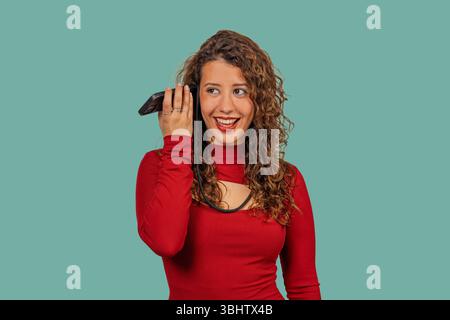 Young woman with curly hair holding smartphone to her ear, listening to a voice message with a curious and happy expression Stock Photo