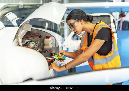 Young woman aircraft mechanic wearing safety glasses and high visibility vest using pliers while working on a small airplane engine inside hangar Stock Photo