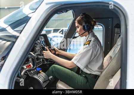 Confident female pilot wearing headset and holding steering wheel while sitting in cockpit of airplane Stock Photo