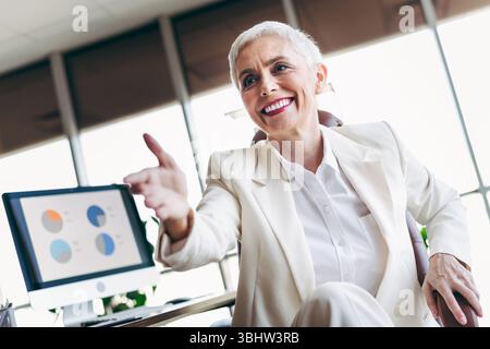 Businesswoman in a white suit extending her hand in a modern office setting with charts on screen. Stock Photo