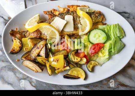 Trout in breadcrumb with lemon, butter and pile of roasted vegetable (aubrgine,cucumber,pepper and zucchini) on white plate on table. Stock Photo