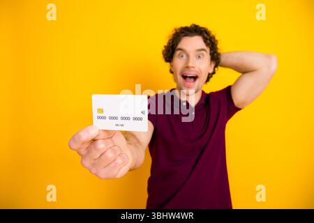 Excited young man in casual burgundy polo holding a credit card on vibrant yellow background, promotional concept Stock Photo