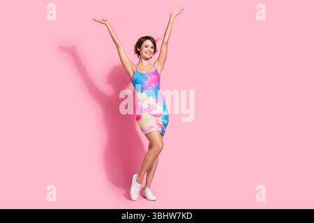 Joyful young woman in colorful summer dress posing with arms raised against pink background Stock Photo