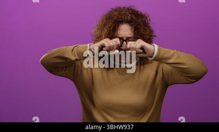 Woman rubbing eyes looking incredulous over bright pink wall wearing glasses brown sweater middle age brunette hispanic isolated setting conveying dis Stock Photo