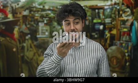 Young hispanic man with beard recording voice message on smartphone in indoor bazar surrounded by eclectic items and wearing striped shirt. Stock Photo