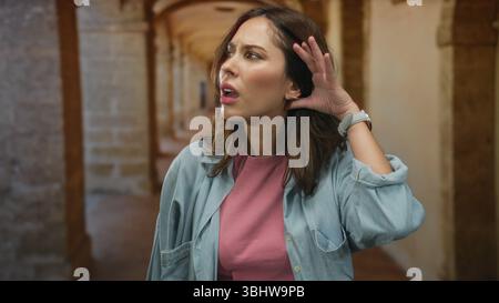 Woman in university hallway listening intently with hand on ear, capturing curiosity and interest in architectural setting. Stock Photo