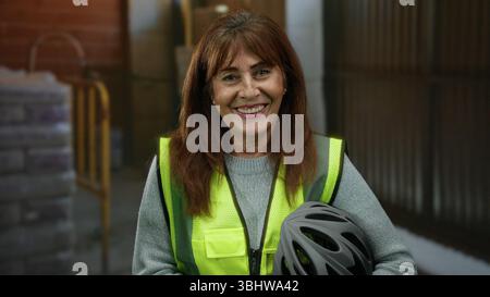 Hispanic senior woman smiling at a construction site indoors, wearing a reflective safety vest and holding a helmet against a background of industrial Stock Photo