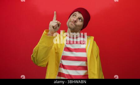 Young hispanic man wearing a yellow raincoat and red beanie makes a thoughtful gesture against a red background suggesting he has an idea. Stock Photo