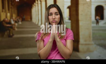 Woman in pink shirt praying with eyes closed in ancient university courtyard. Stock Photo