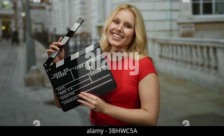 Woman holding clapperboard wearing red shirt smiling on city street outdoors showing filmmaking process. Stock Photo