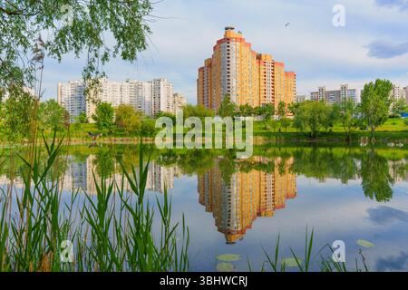 Wide-angle view of modern residential buildings reflected in a serene lake, surrounded by greenery under a partly cloudy sky. Stock Photo