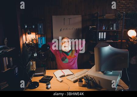 Elderly IT Specialist Relaxing in a Cozy Home Office Environment Working on a Computer Late in the Evening Stock Photo