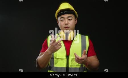 Man wearing safety vest and hard hat gestures playfully against a black background, showcasing confidence and safety awareness on site. Stock Photo